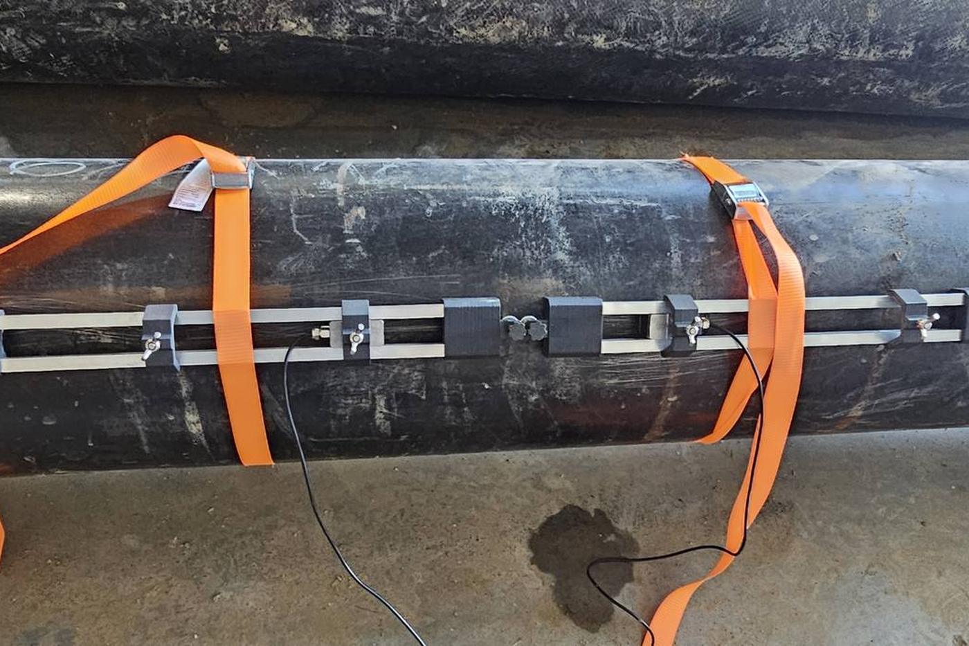 Clamp-on ultrasonic transducer rail strapped to a large-diameter black steel pipe with orange ratchet straps